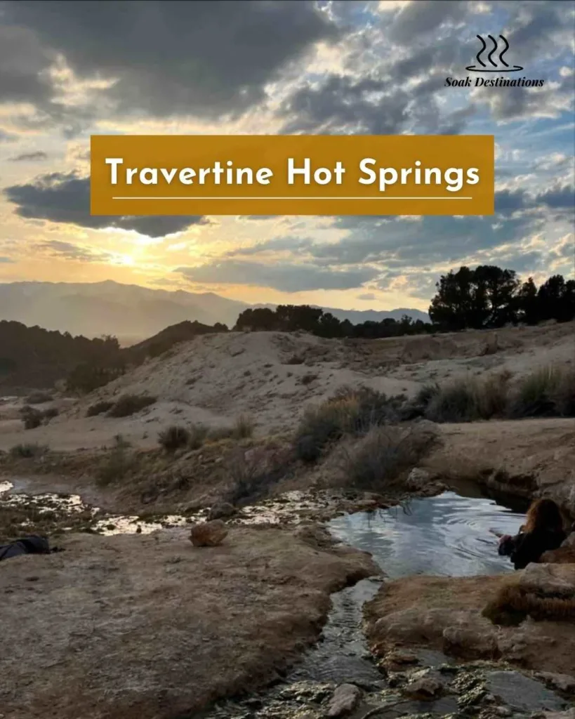 A person relaxes in the Travertine Hot Springs while watching the sunset over distant desert mountains.