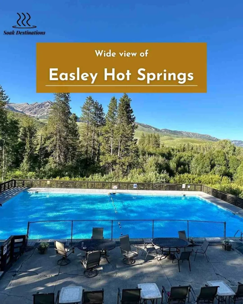 Wide view of Easley Hot Springs pool with patio seating, forest scenery, and clear blue sky