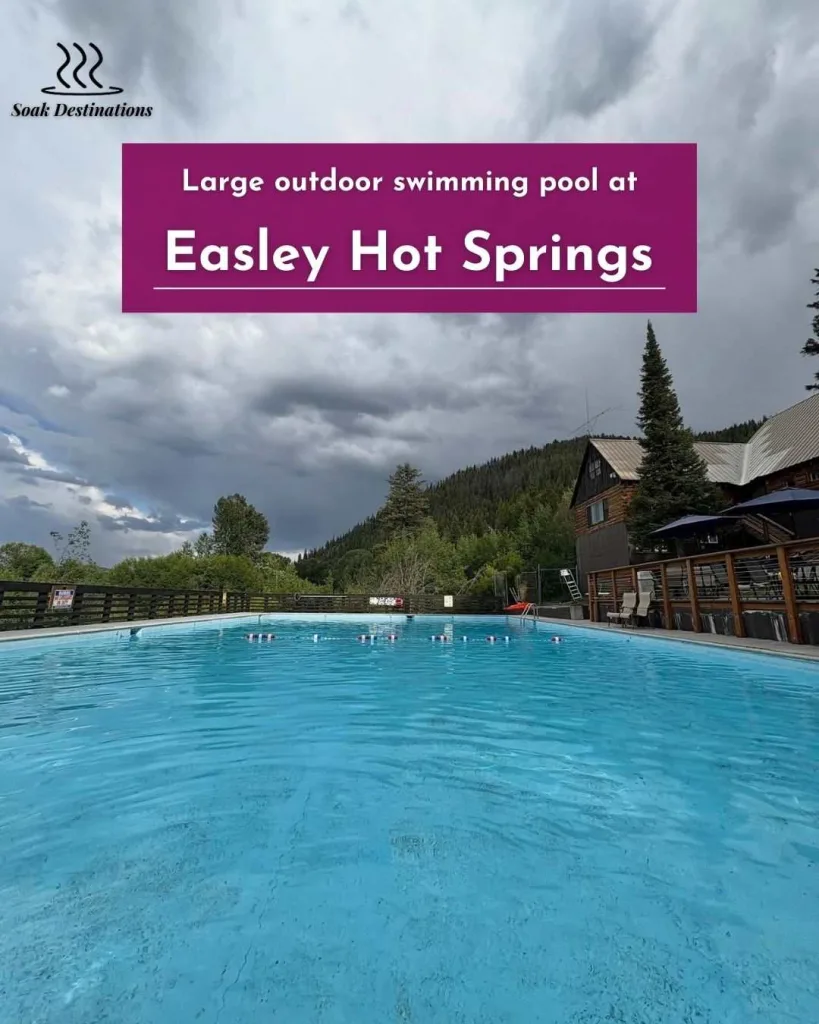 Large outdoor swimming pool at Easley Hot Springs surrounded by mountains, cabins, and dramatic cloudy skies.
