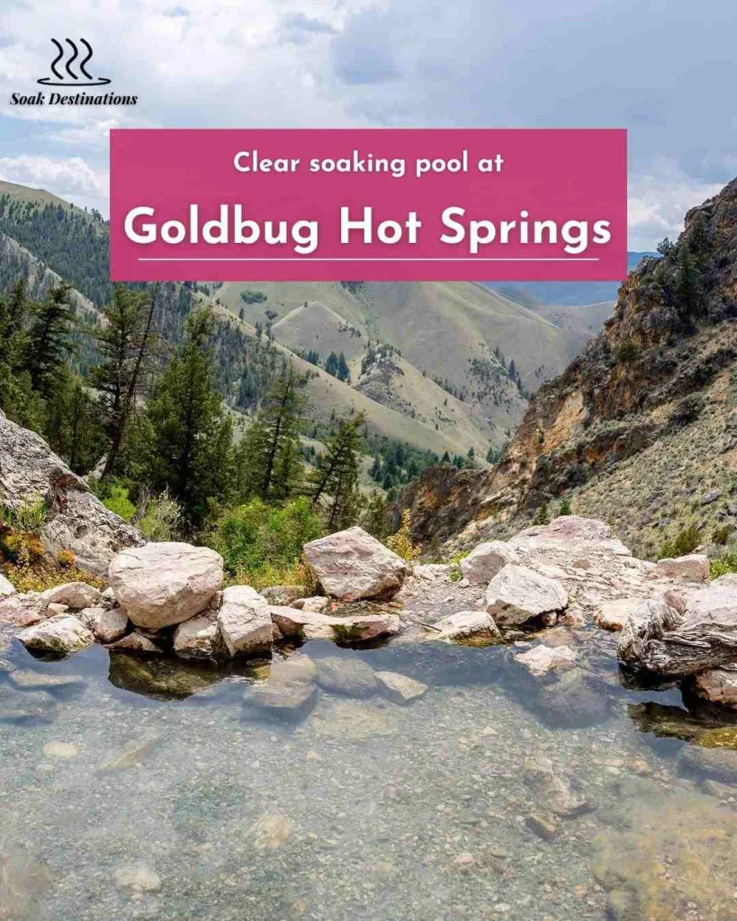 Clear soaking pool at Goldbug Hot Springs overlooking green mountains, rocky slopes, and cloudy skies.